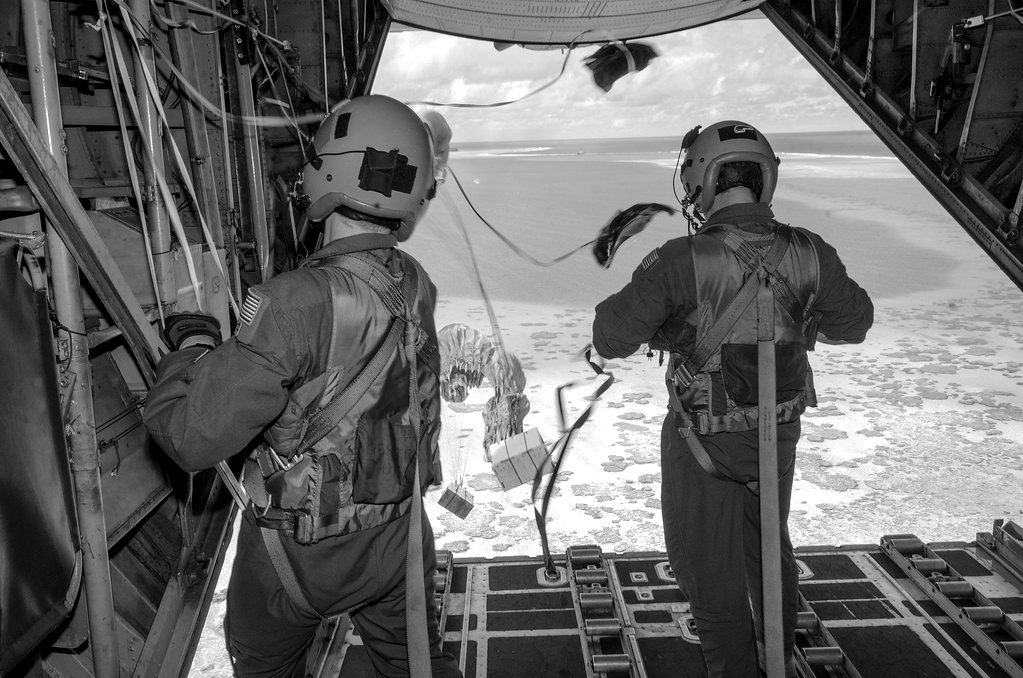 Crew watching supply bundles parachute from an open aircraft ramp over the sea