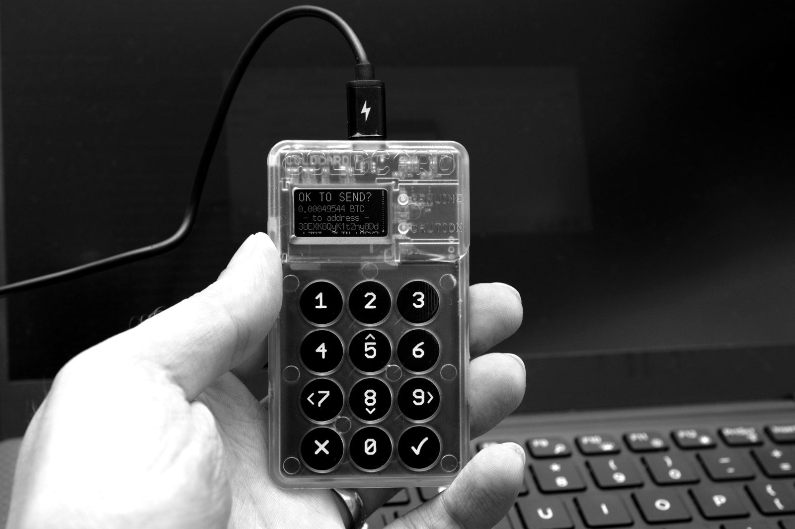 A hand holding a transparent hardware wallet with a numeric keypad, connected by USB